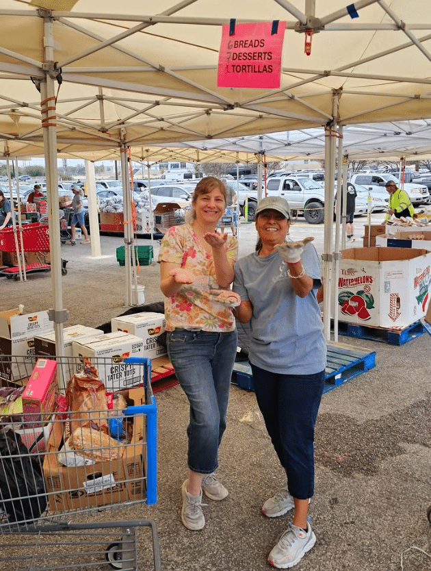 Two volunteers smiling while serving at a Coker Methodist Church food pantry outreach under outdoor tents in San Antonio.